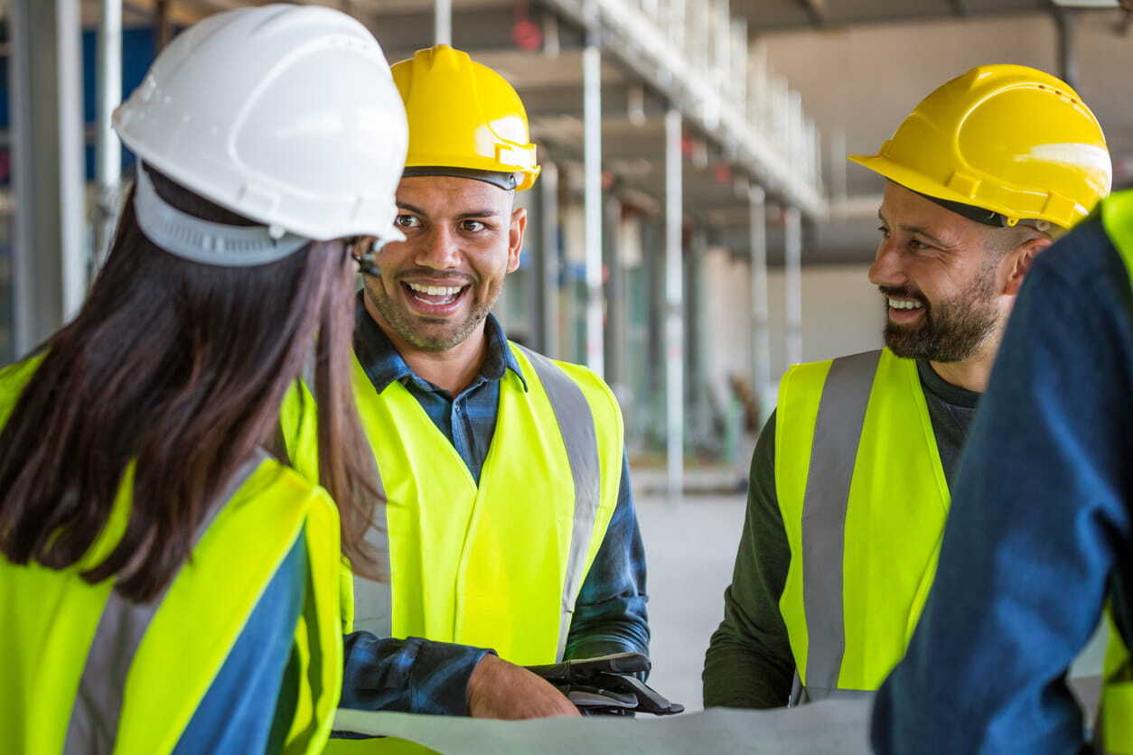 Man smiling in hard hat and high vis vest holding a tablet and chatting with two colleagues on a construction site_min