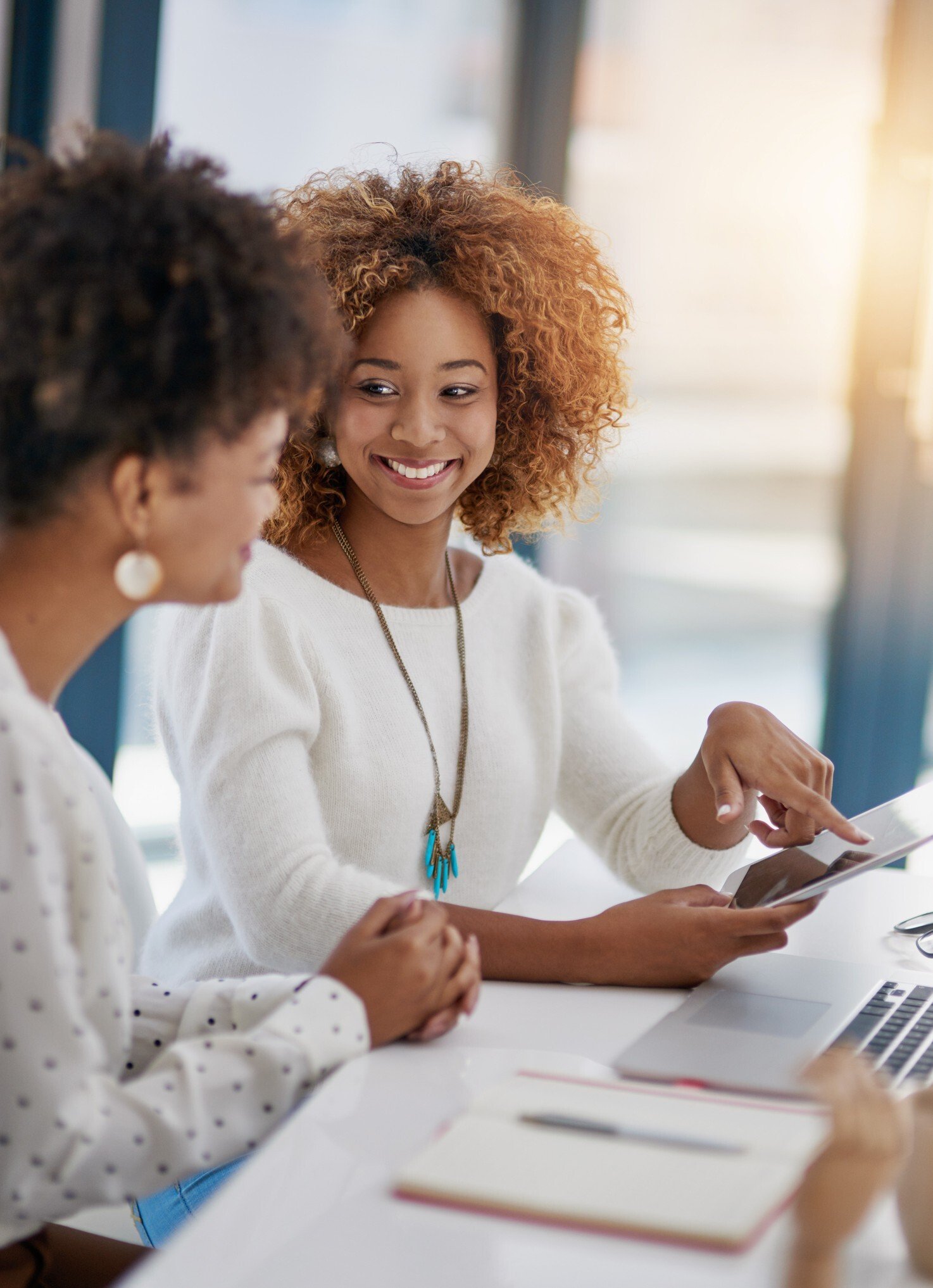 Two female workers chatting over a laptop and tablet in an office environment