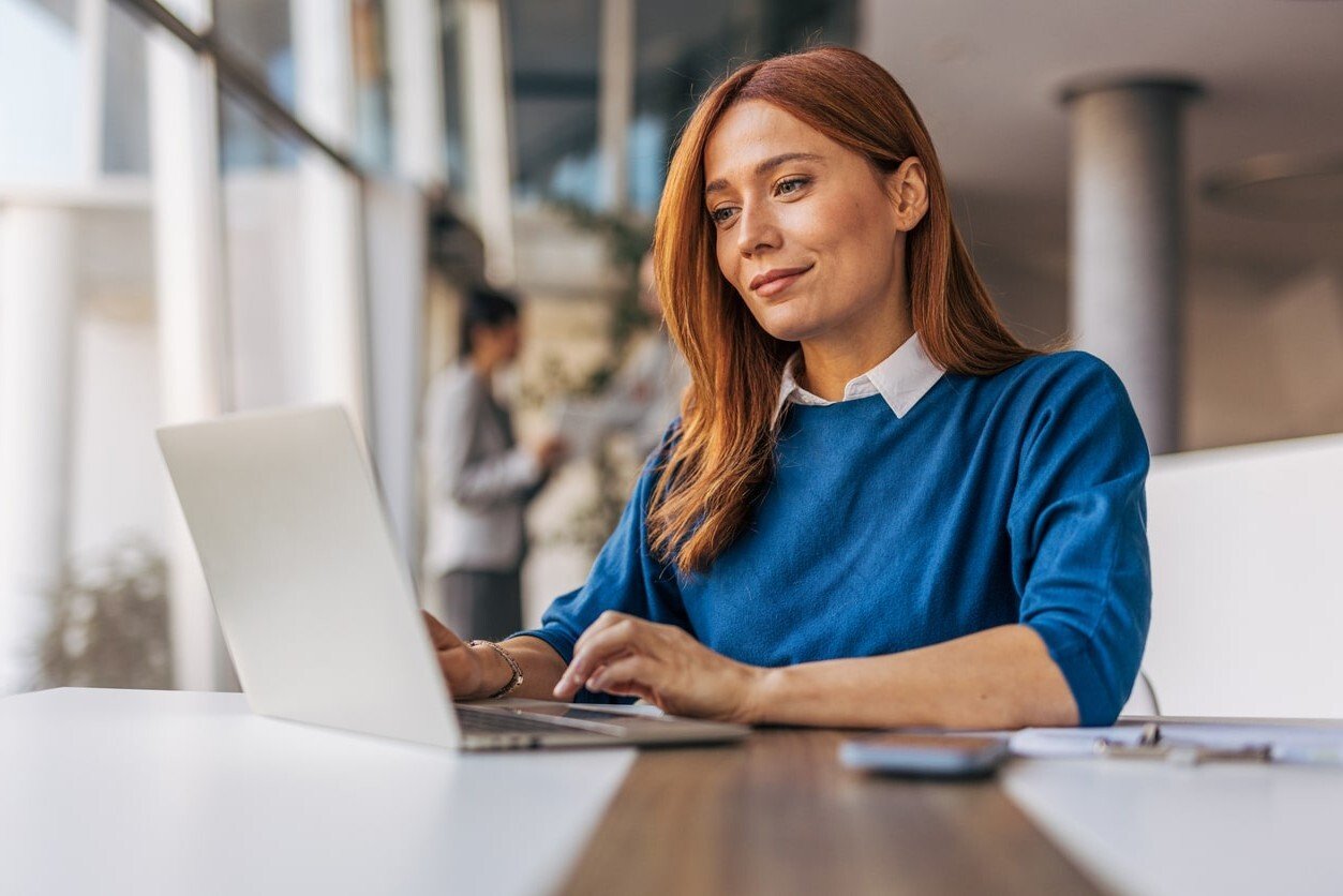 Young woman with red hair, wearing blue top sits at desk with open laptop_facing left